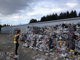 A person stands in front of a large amount of waste from a baler or a compactor. There are trees in the background.