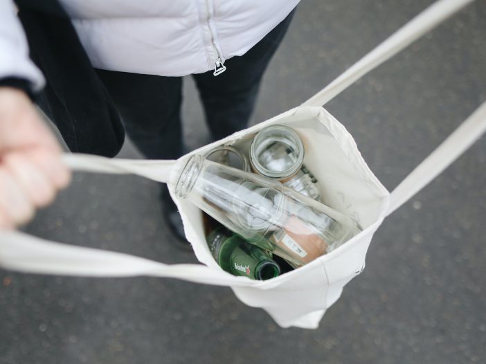 5 ways to improve your recycling. A person is holding a white reusable shopping bag which is filled with empty plastic and glass bottles/containers.
