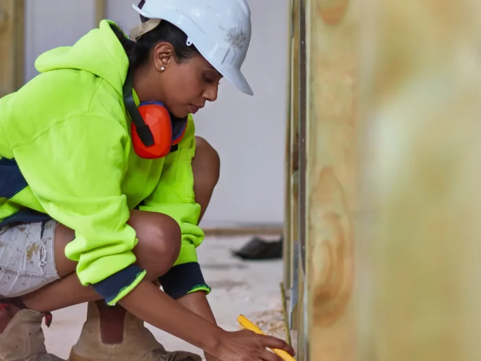 Training for a Certificate in Millwrighting. A female construction worker is taking a measurement. She is wearing a high-vis jacket and a white helmet.