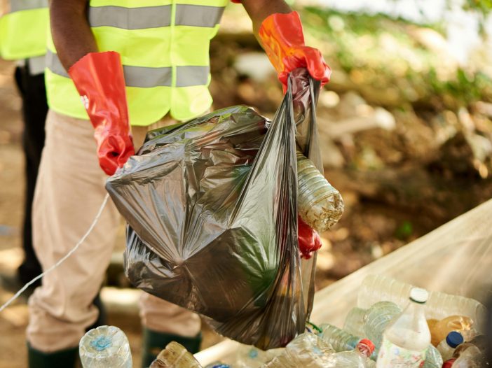 How Your Business Can Reduce Plastic Consumption. A person is wearing red gloves and a high-vis vest, holding a bag of plastic waste. They are standing behind a big pile of plastic waste.